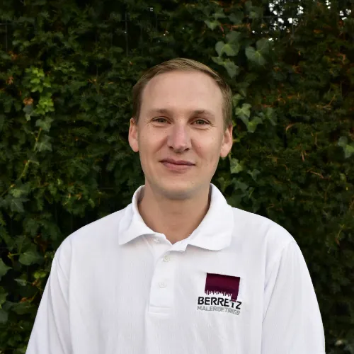 Junger Mann mit kurzem Haar und weißem Poloshirt vor grünem Laub, auf dem Hemd steht Berretz Malerbetrieb.