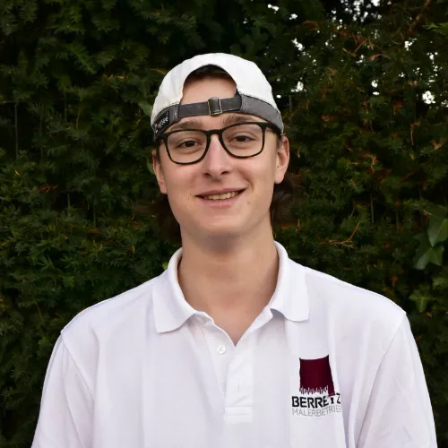 Junger Mann mit Brille, weißer Baseballmütze rückwärts und weißem Polohemd mit Firmenlogo vor grünem Laub.