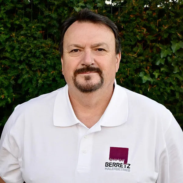 Mann in weißem Poloshirt mit Firmenlogo Berretz Malerbetrieb vor grünem Laub.