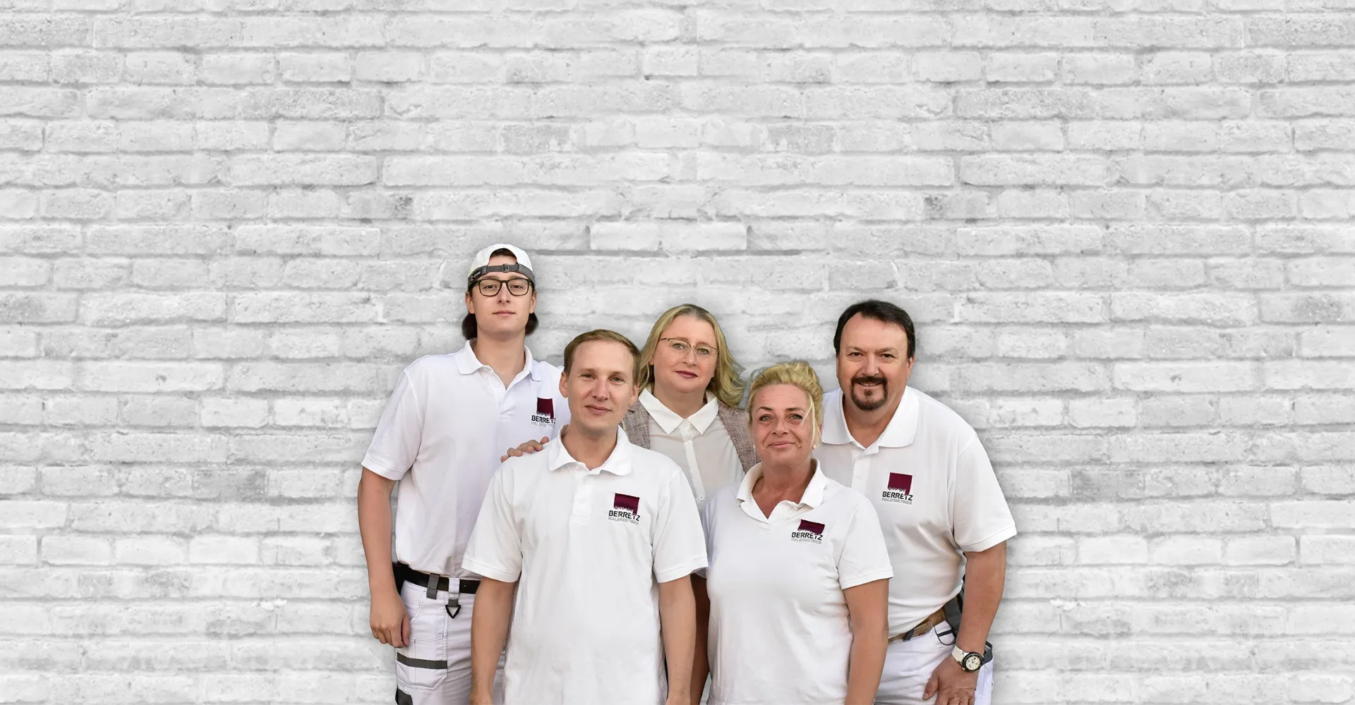 Fünf Personen vor einer weißen Ziegelsteinwand, vier tragen weiße Poloshirts mit dem Schriftzug Berretz Malerbetrieb, eine Frau in der Mitte trägt eine helle Bluse und Jacke.